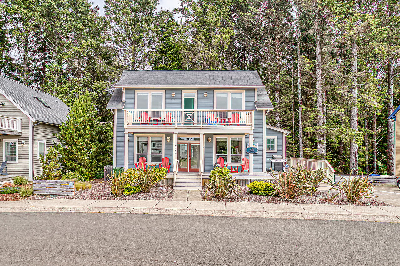 A charming blue coastal-style home nestled among tall pines, featuring bright red outdoor seating and welcoming balconies for relaxing in the fresh forest air.